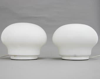 May include: Two white mushroom-shaped table lamps with a round base. The lamps are on a gray surface.