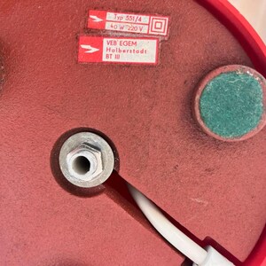 May include: Close-up of the underside of a red lamp base. The base has two green felt pads and a central metal fitting. Two white labels with black text are visible, including "Typ 551/4" and "VEB EGEM Holberstadt BT III". A white power cord extends from the base.
