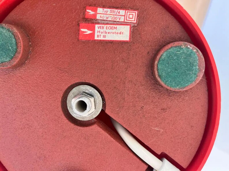 May include: Close-up of the underside of a red lamp base. The base has two green felt pads and a central metal fitting. Two white labels with black text are visible, including "Typ 551/4" and "VEB EGEM Holberstadt BT III". A white power cord extends from the base.