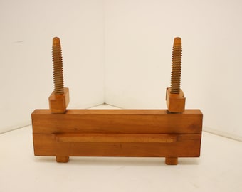 May include: A wooden book press with two threaded spindles. The press is made of light brown wood and has a rectangular base with two feet. The spindles are turned from wood and are used to apply pressure.