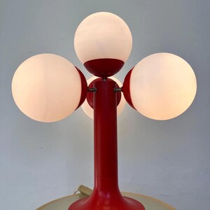 May include: A red table lamp with four white globe-shaped shades. The lamp has a central red stem and base, with the shades arranged around the top. The lamp is on a light-colored table.