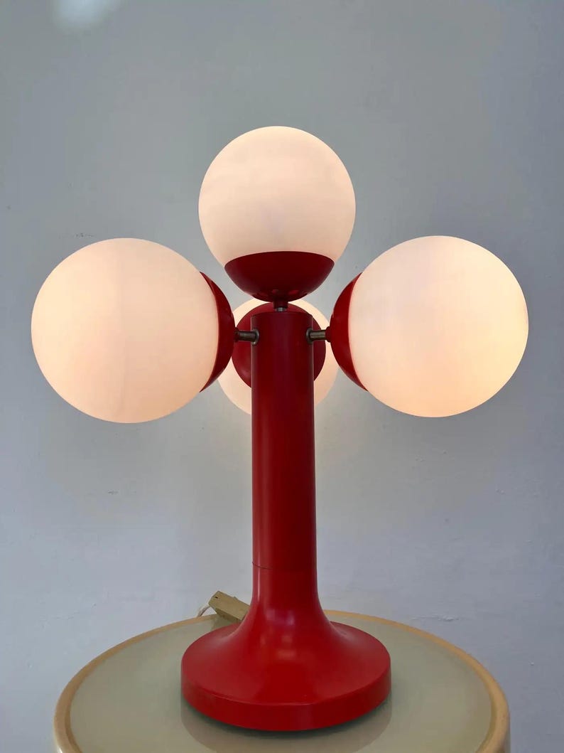 May include: A red table lamp with four white globe-shaped shades. The lamp has a central red stem and base, with the shades arranged around the top. The lamp is on a light-colored table.