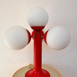May include: A red table lamp with three white globe shades. The lamp has a round base and a central stem. The shades are evenly spaced around the stem. The lamp is a retro design, perfect for a living room or bedroom.