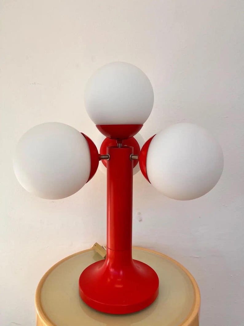 May include: A red table lamp with three white globe shades. The lamp has a round base and a central stem. The shades are evenly spaced around the stem. The lamp is a retro design, perfect for a living room or bedroom.