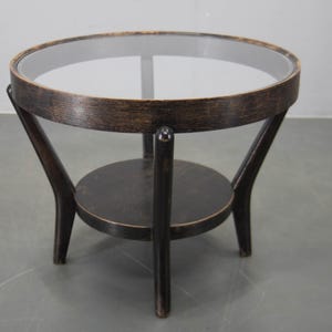May include: A small, round side table with a dark wood frame and a clear glass top. The table has a lower shelf and three curved legs. The table is approximately 61 cm in diameter and 51 cm tall.
