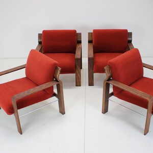 May include: Four mid-century modern armchairs. Each chair has a red cushion and backrest, with a wooden frame and armrests. The chairs have a minimalist design and are arranged in a group.