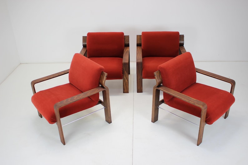 May include: Four mid-century modern armchairs. Each chair has a red cushion and backrest, with a wooden frame and armrests. The chairs have a minimalist design and are arranged in a group.
