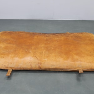 Könnte beinhalten: Rechteckiges, abgenutztes Lederkissen in Brauntönen. Das Kissen hat eine leicht gebogene Form und sichtbare Nähte an den Kanten. Zwei Lederriemen sind unten befestigt. Das Kissen scheint Vintage zu sein.