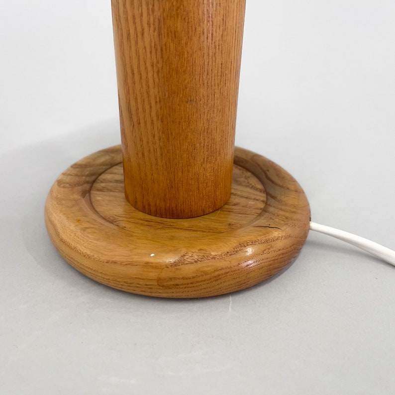 Puede incluir: Una l&aacute;mpara de mesa de madera con una base cil&iacute;ndrica y una base redonda de madera. La l&aacute;mpara tiene un cable blanco.