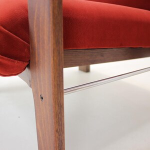 May include: Close-up of a mid-century modern chair. The chair features a vibrant red velvet seat and backrest, complemented by a dark wood frame. A polished metal bar adds a sleek accent. The design showcases clean lines and a retro aesthetic.