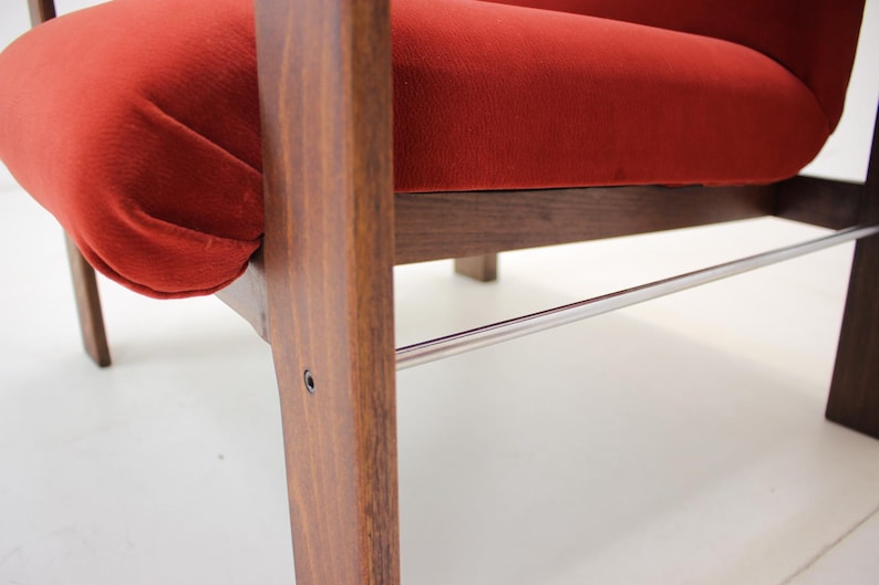 May include: Close-up of a mid-century modern chair. The chair features a vibrant red velvet seat and backrest, complemented by a dark wood frame. A polished metal bar adds a sleek accent. The design showcases clean lines and a retro aesthetic.