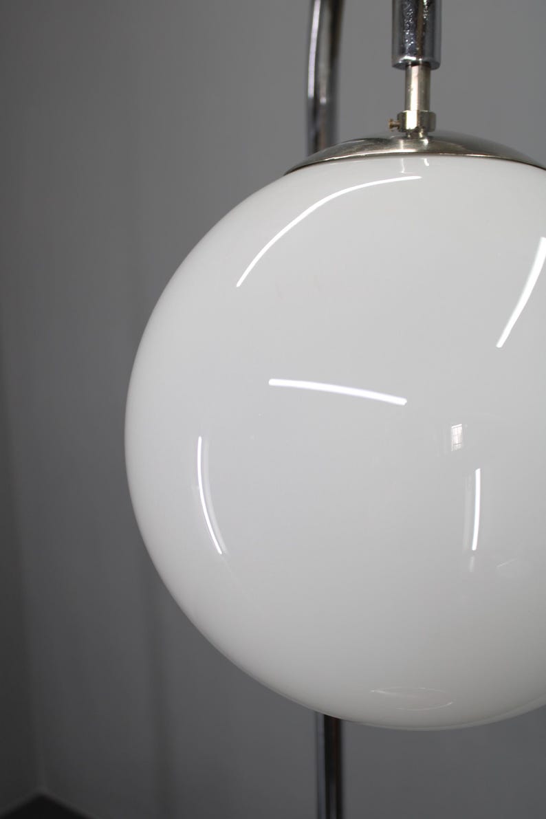 May include: Close-up of a white, spherical lamp shade with a glossy finish. The shade is part of a chrome and silver floor lamp. The lamp is against a gray background.