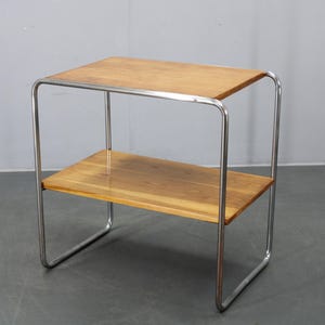May include: A small side table with two wooden shelves and a chrome frame. The rectangular shelves are made of light-colored wood. The frame is made of curved, tubular chrome. The table is a simple, modern design.