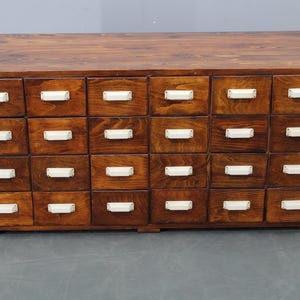 May include: A wooden cabinet with twenty-four drawers. The cabinet is made of dark brown wood with a natural grain. Each drawer has a white rectangular handle. The cabinet is a storage unit.