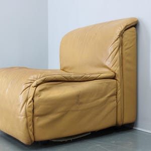 May include: A tan leather armchair with a unique, curved design. The chair has a tufted back and seat, and the leather is slightly distressed.