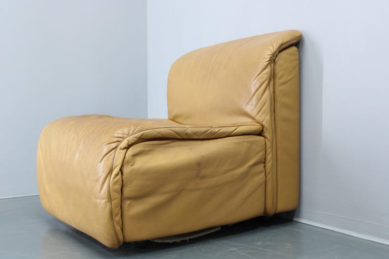 May include: A tan leather armchair with a unique, curved design. The chair has a tufted back and seat, and the leather is slightly distressed.