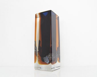 May include: A rectangular glass vase with a clear base and a dark, abstract design. The design features vertical bands of black, orange, and white, with speckled accents. The top of the vase has a dark blue hue.