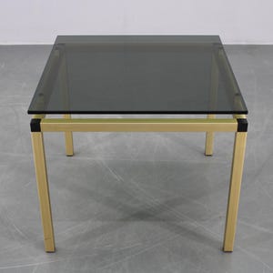 May include: A square coffee table with a smoked glass top and a gold-coloured metal frame. The table has a minimalist design with a sleek, modern aesthetic. The legs are straight and angled slightly outward.