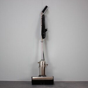 May include: A vintage upright vacuum cleaner with a silver metal body and a black handle. The vacuum has a rectangular brush head and a black power cord. The design is simple and functional, with a focus on cleaning.