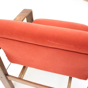 May include: Close-up of a mid-century modern chair with a burnt orange velvet seat and backrest. The wooden frame has a warm, natural finish. The chair's design features curved armrests and a slatted base.