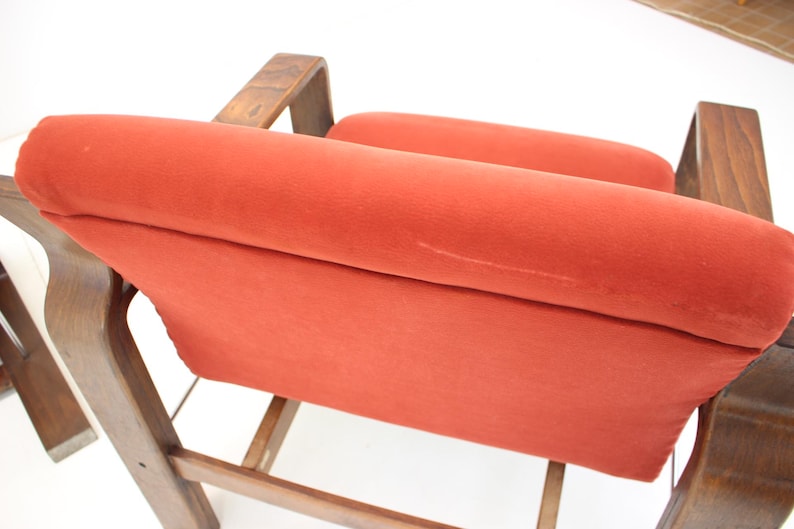 May include: Close-up of a mid-century modern chair with a burnt orange velvet seat and backrest. The wooden frame has a warm, natural finish. The chair's design features curved armrests and a slatted base.