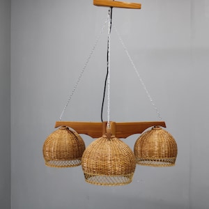 Può includere: Lampada a sospensione a tre luci con paralumi in rattan intrecciato. L'apparecchio ha una struttura in legno ed è sospeso da catene e un cavo nero. I paralumi sono di un colore caldo e naturale, e il design ricorda lo stile moderno della metà del secolo.