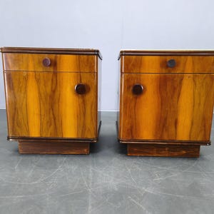 May include: Two matching wooden nightstands with a warm, striped wood grain pattern. Each nightstand has a drawer with a round knob and a circular handle on the door. The nightstands are set on a dark brown base.