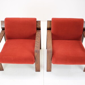 May include: Two mid-century modern armchairs. Each chair features a vibrant red seat and backrest, complemented by a dark wood frame and armrests. The chairs are set against a plain white background, highlighting their design.