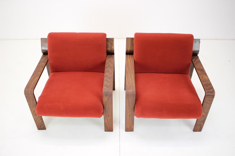 May include: Two mid-century modern armchairs. Each chair features a vibrant red seat and backrest, complemented by a dark wood frame and armrests. The chairs are set against a plain white background, highlighting their design.