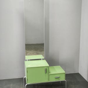 Peut inclure: Un meuble de toilette vert clair doté d'un grand miroir rectangulaire fixé sur une base comprenant une porte de placard et un tiroir. L'ensemble repose sur un cadre en métal argenté à quatre pieds et comporte des poignées argentées.