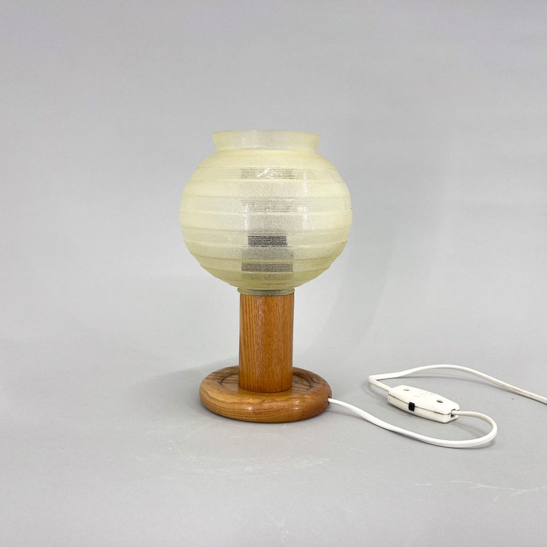 Puede incluir: Una pantalla de l&aacute;mpara blanca con nervaduras en forma de globo se asienta sobre una base de madera con un cable y un interruptor. La pantalla est&aacute; hecha de un material transl&uacute;cido y la base es de color marr&oacute;n c&aacute;lido.