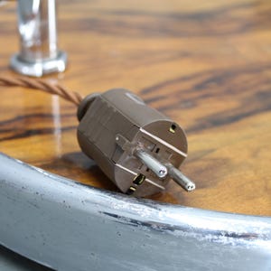 May include: Close-up of a brown electrical plug with two prongs. The plug is set against a wooden surface with a chrome metal rim. A brown cord extends from the plug. The background is blurred.