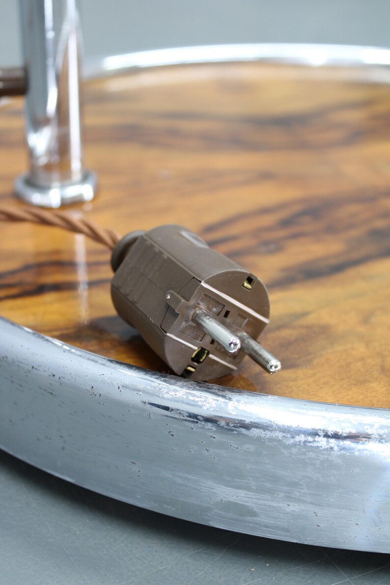 May include: Close-up of a brown electrical plug with two prongs. The plug is set against a wooden surface with a chrome metal rim. A brown cord extends from the plug. The background is blurred.