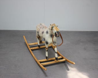 May include: A vintage-style rocking horse with a spotted white and gray body, black hooves, and a brown leather bridle. The horse is mounted on a wooden rocker with a natural finish, creating a classic toy aesthetic.