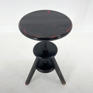 May include: Black adjustable height stool with a round seat and three legs. The seat has red paint splatter.