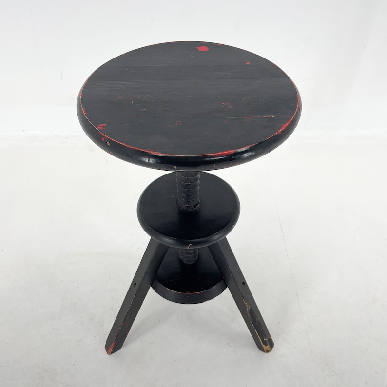 May include: Black adjustable height stool with a round seat and three legs. The seat has red paint splatter.