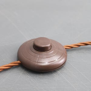 May include: A brown, circular foot switch with a raised button in the center. The switch has concentric circle detailing and is connected to a brown, braided electrical cord. The background is a solid gray.