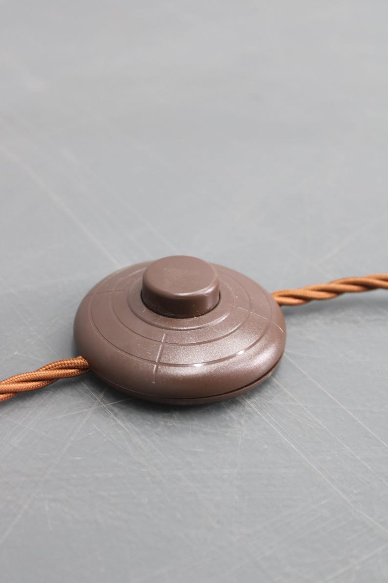 May include: A brown, circular foot switch with a raised button in the center. The switch has concentric circle detailing and is connected to a brown, braided electrical cord. The background is a solid gray.