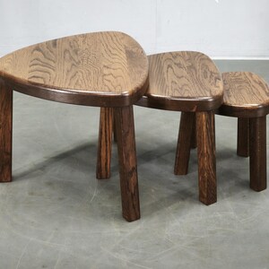 May include: A set of three nesting tables crafted from dark brown wood with a visible grain pattern. Each table features a smooth, rounded triangular top surface supported by three sturdy, straight legs, designed to stack neatly within one another.
