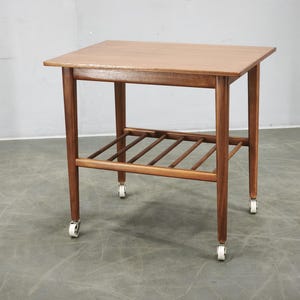 May include: A rectangular wooden side table with a smooth top surface and four straight legs. The table features a lower slatted shelf for storage and is supported by four small white swivel casters attached to the base of each wooden leg.