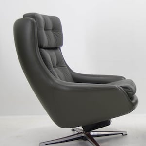 Op de afbeelding: Een donkergrijze, moderne fauteuil uit het midden van de eeuw met een hoge rugleuning en geïntegreerde hoofdsteun. De stoel heeft een gecapitonneerd ontwerp op de rug en hoofdsteun, met een gebogen zitting en armleuningen. Hij draait op een chroom en houten voet.