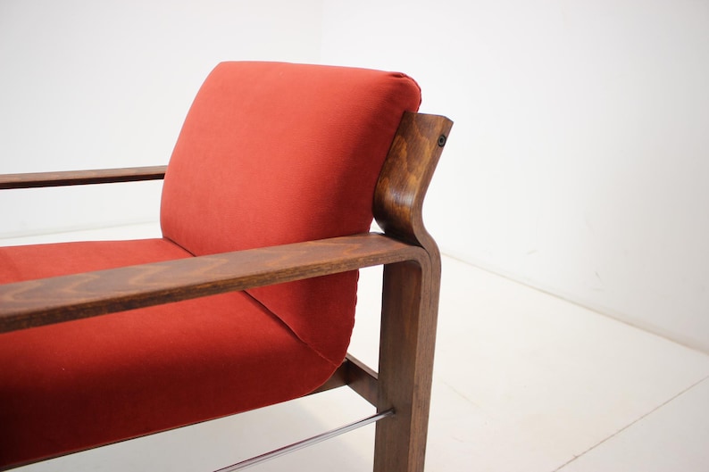 May include: A mid-century modern armchair with a vibrant red cushion and dark wood frame. The chair features a curved backrest and armrests, with a sleek, minimalist design. The frame is made of dark wood, and the cushion is a solid red color.