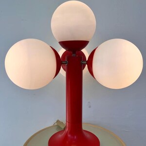 May include: A red table lamp with three spherical white shades. The lamp has a cylindrical red stem and a round base. The shades are evenly spaced around the stem, creating a unique lighting fixture.