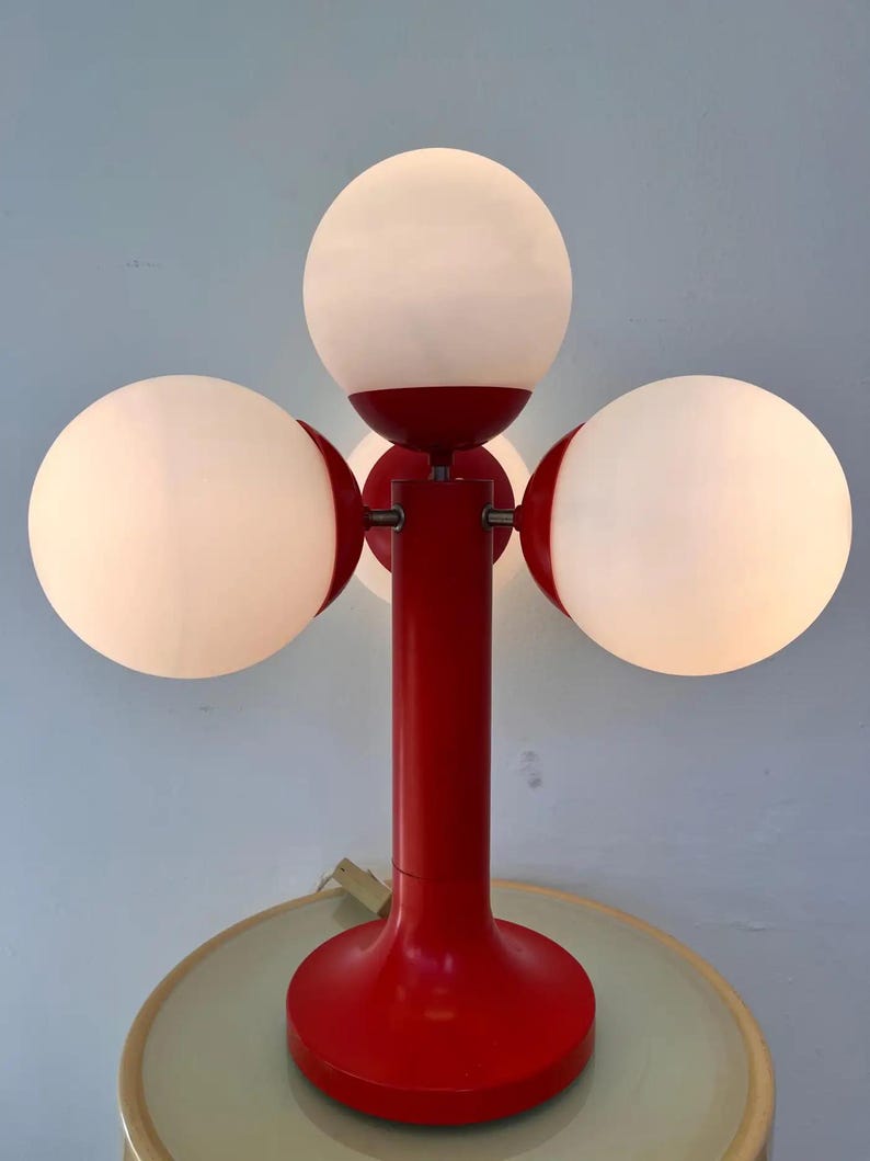 May include: A red table lamp with three spherical white shades. The lamp has a cylindrical red stem and a round base. The shades are evenly spaced around the stem, creating a unique lighting fixture.