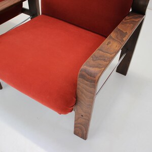 May include: Close-up of a mid-century modern chair with a vibrant orange seat and dark wood frame. The chair features a curved armrest and sturdy legs, showcasing a blend of comfort and style. The design is reminiscent of classic furniture.