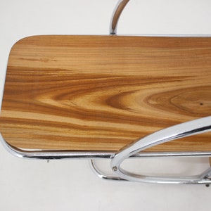 May include: A chrome and wood serving cart with two shelves. The top shelf is made of wood with a natural grain pattern. The cart has a curved design and is made of polished chrome.