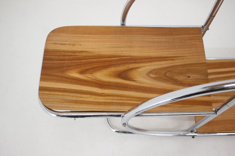 May include: A chrome and wood serving cart with two shelves. The top shelf is made of wood with a natural grain pattern. The cart has a curved design and is made of polished chrome.