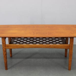 May include: A mid-century modern coffee table with a warm wood finish. The rectangular tabletop has rounded corners and sits atop four tapered legs. A shelf with a perforated design is located below the tabletop. The table measures approximately 48 inches long.