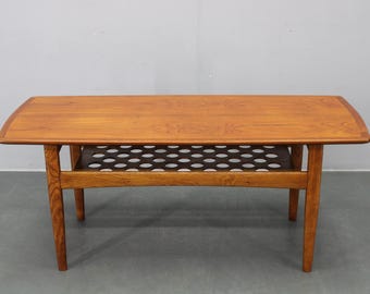 May include: A mid-century modern coffee table with a warm wood finish. The rectangular tabletop has rounded corners and sits atop four tapered legs. A shelf with a perforated design is located below the tabletop. The table measures approximately 48 inches long.
