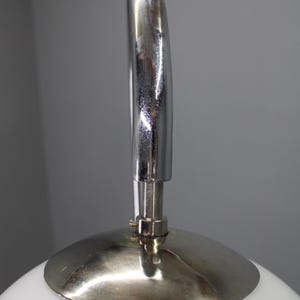 May include: Close-up of a white, rounded lamp with a silver metal handle and base. The handle arches over the top of the lamp, and the base is a circular disc. The lamp has a modern, minimalist design.